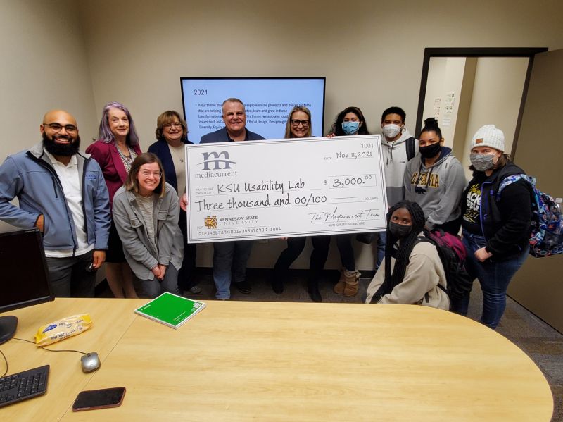 A diverse group of students, teachers, and Mediacurrent team members hold up a giant check for the KSU usability lab